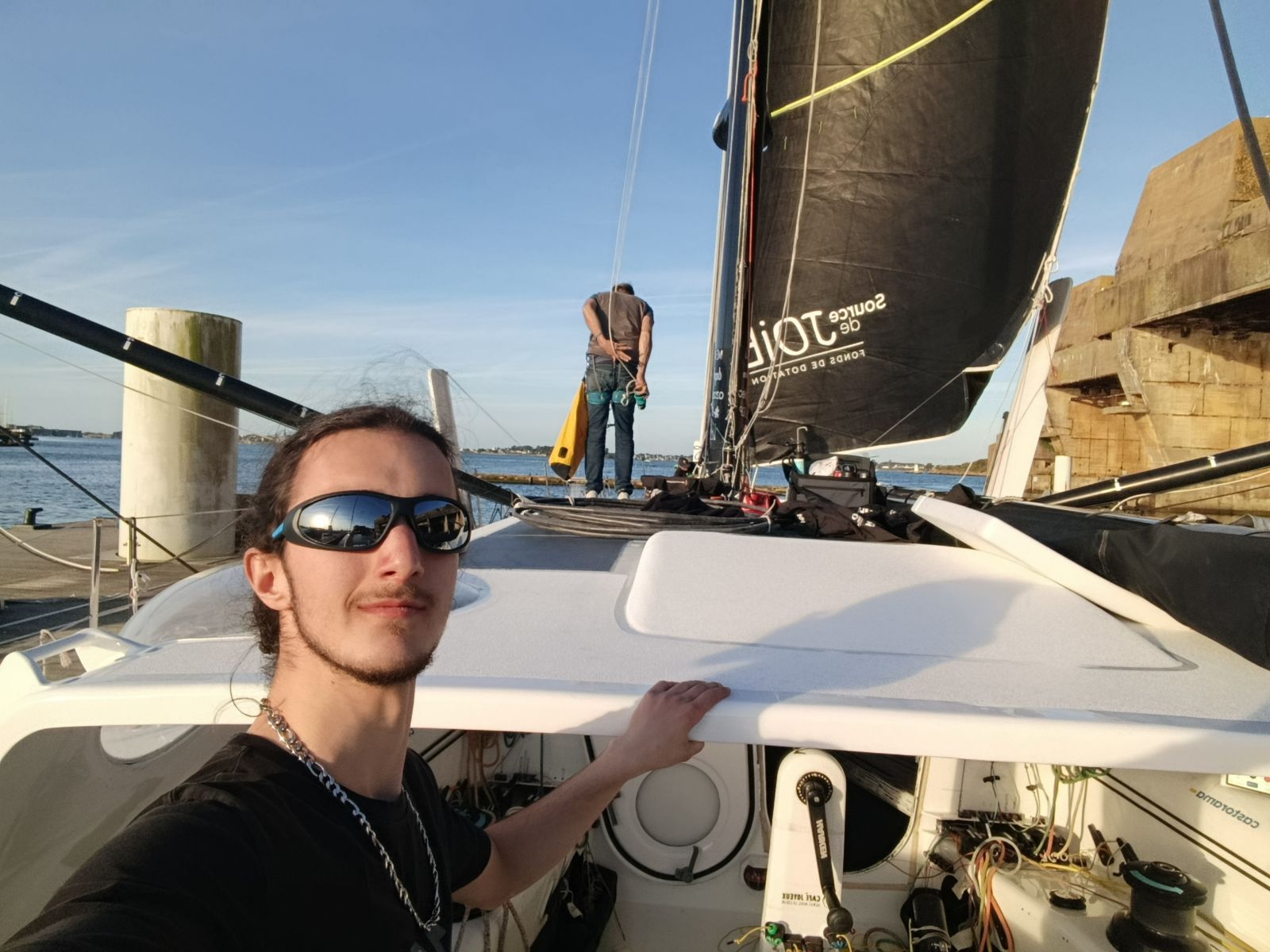 Jules Poulain à quai devant un voilier de course gréé, un équipier travaillant sur le pont à l’arrière-plan.