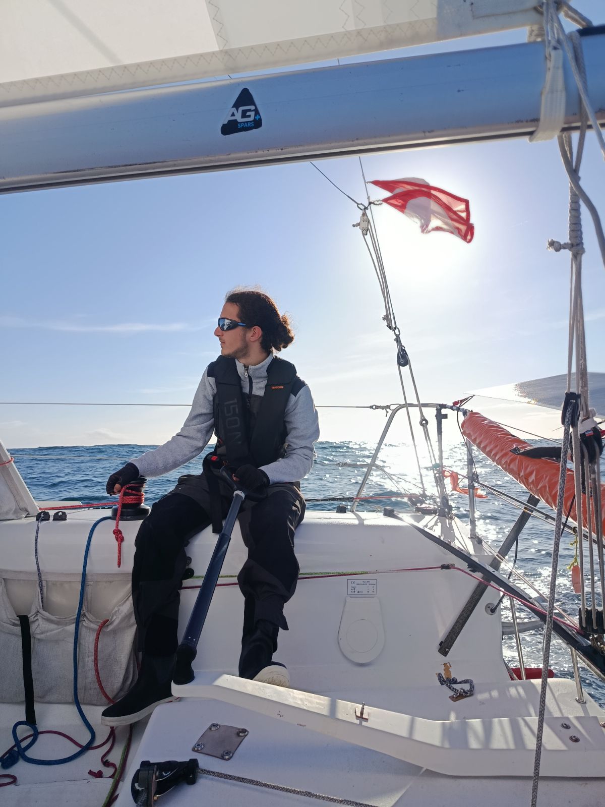 Jules Poulain à la barre franche d’un voilier de course, en gilet de sauvetage, au large par grand soleil.