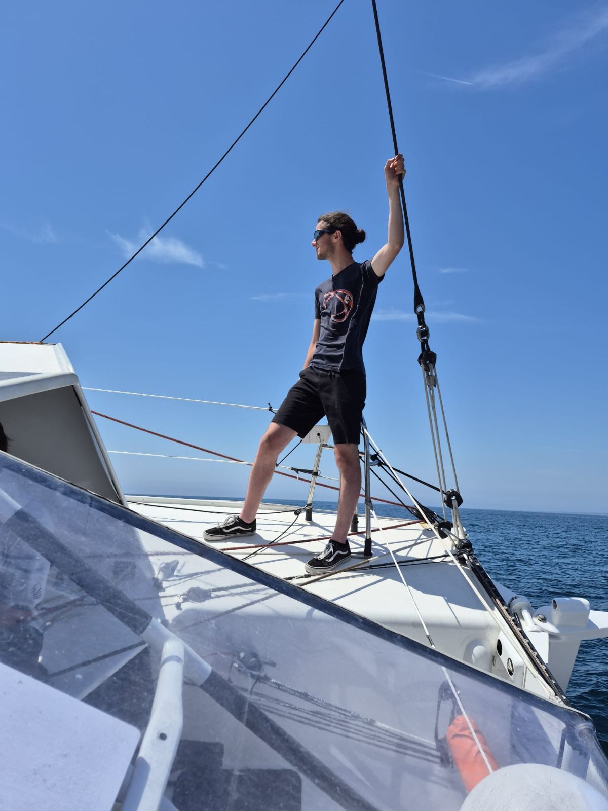 Jules Poulain debout sur le pont d’un IMOCA, une main sur une drisse, par mer calme et grand soleil.