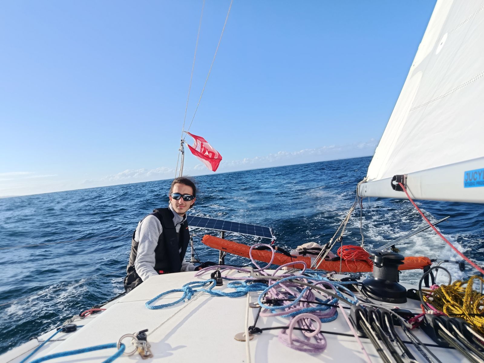 Jules Poulain à bord d’un voilier de course au large, assis au rappel dans le cockpit, mer bleue et ciel dégagé.