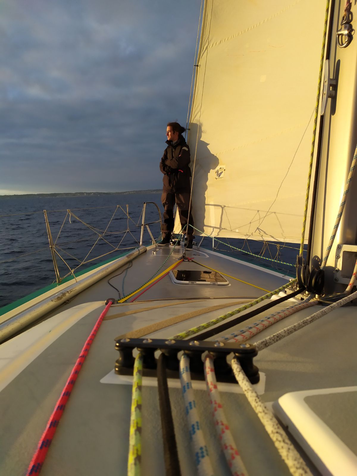 Jules Poulain debout à l’avant d’un voilier de course au coucher du soleil, bouts colorés sur le pont au premier plan.