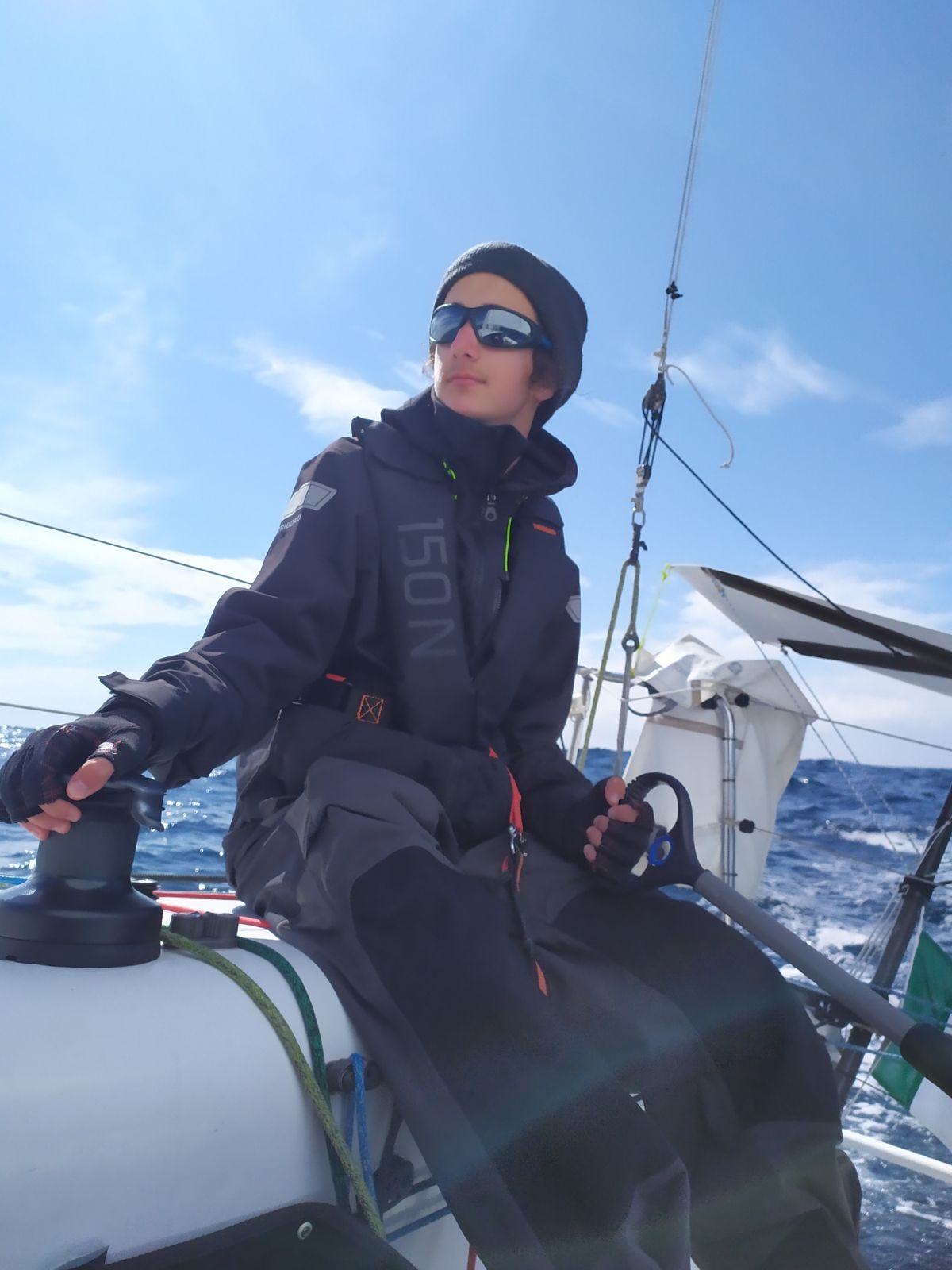 Jules Poulain en ciré et bonnet, barre en main près d’un winch, par mer formée.